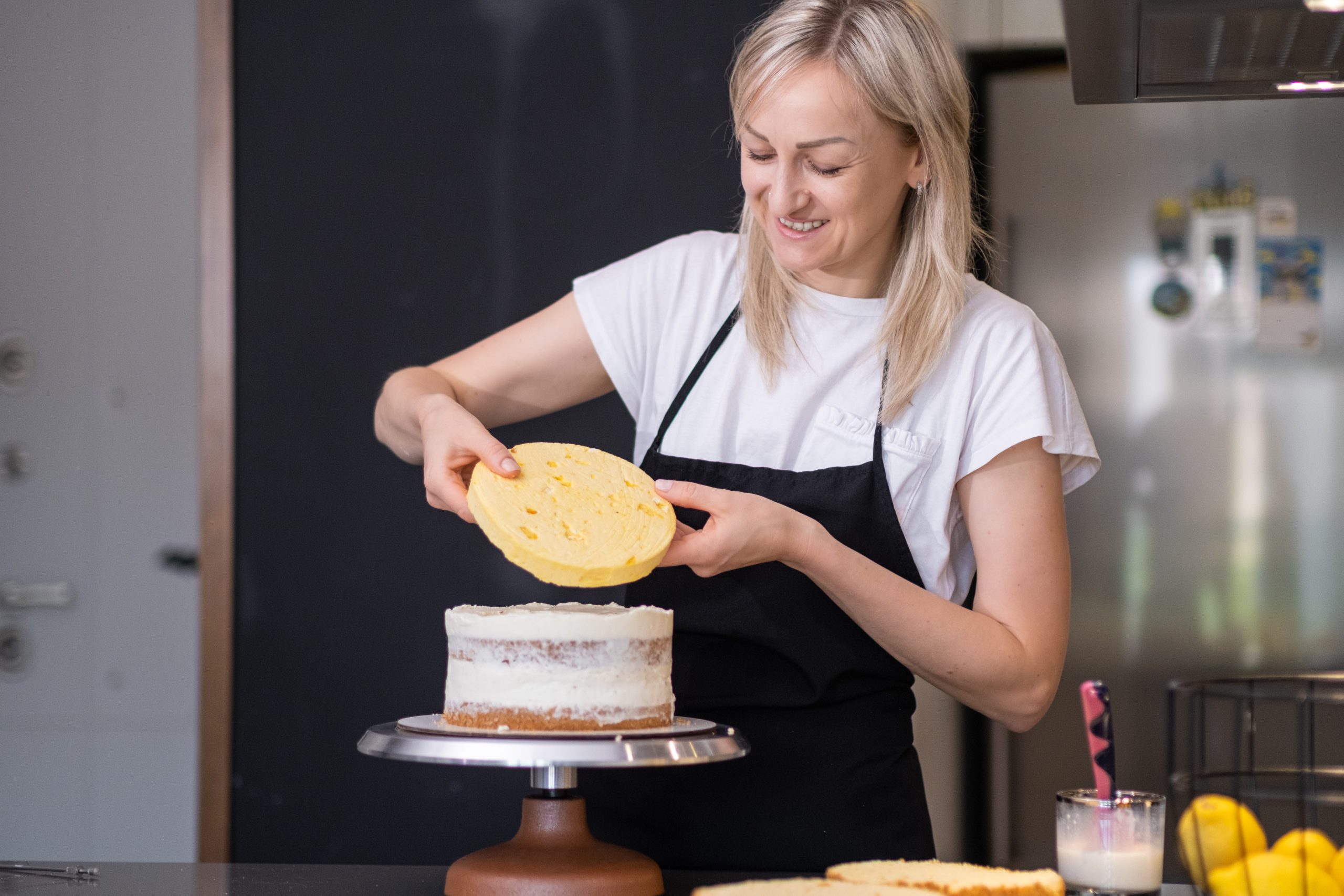 Satisfied blonde woman connecting fresh cake layers smeared with sweet cream smiling happy female confectioner in black apron standing at table making delicious custom pastry